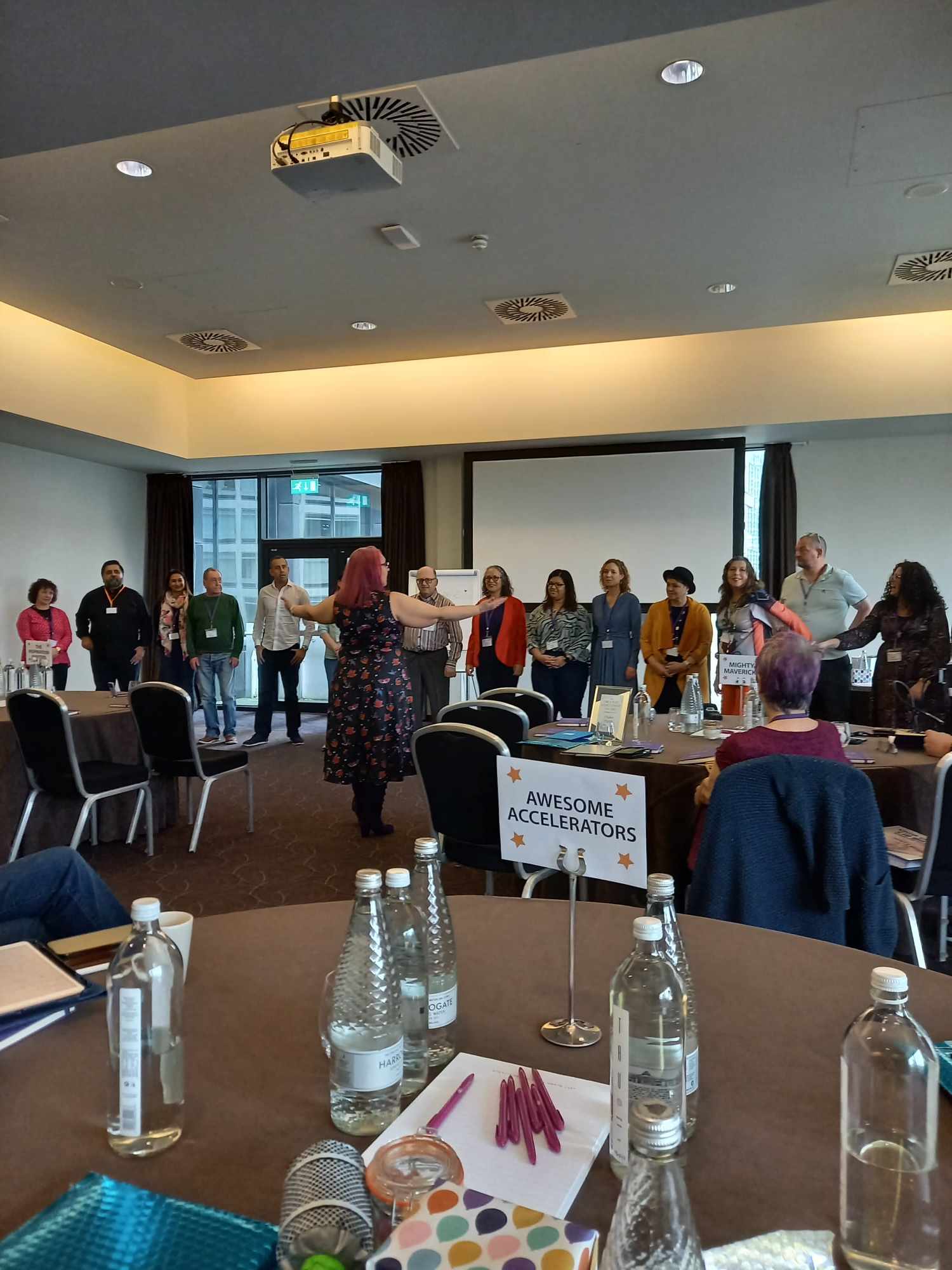 Picture of different people standing in a row att he front of a room with Pollie Standing in front facing them with her arms open, Pollie is wearing a blue dress with red pattern and pink shoulderlength hair 