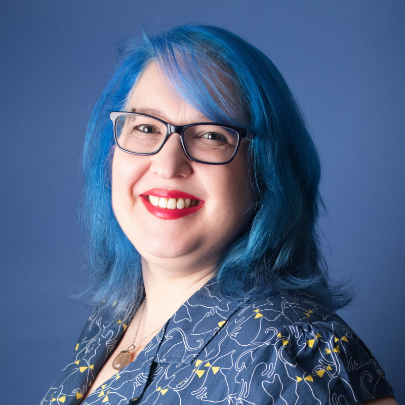 woman with blue hair and glases smiling at the camera wearing a blue dress with yellow detailing 